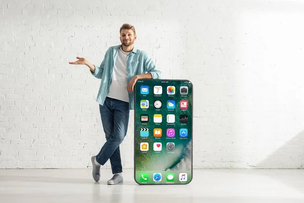 KYIV, UKRAINE - FEVEREIRO 21, 2020: Bonito homem sorrindo enquanto mostra gesto de dubium perto de grande modelo de smartphone com tela de iphone — Fotografia de Stock