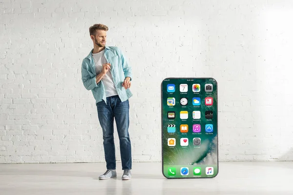 KYIV, UCRANIA - 21 de febrero de 2020: Hombre asustado mirando gran modelo de teléfono inteligente con pantalla de iphone en casa - foto de stock