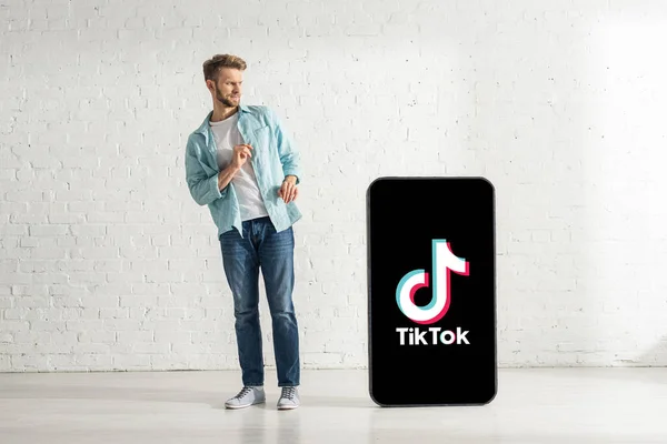 KYIV, UKRAINE - FEBRUARY 21, 2020: Confused man standing near big model of smartphone with TikTok app at home — Stock Photo