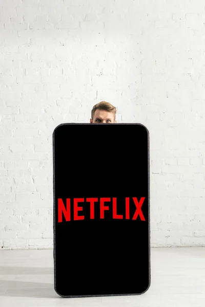 KYIV, UCRANIA - 21 de febrero de 2020: Hombre mirando a la cámara cerca de un gran modelo de teléfono inteligente con aplicación netflix en casa — Stock Photo