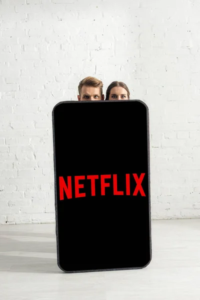 KYIV, UCRANIA - 21 de febrero de 2020: Pareja mirando a la cámara cerca de un enorme modelo de teléfono inteligente con aplicación netflix en casa - foto de stock