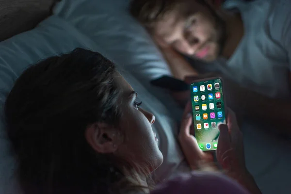 KYIV, UKRAINE - FEBRUARY 21, 2020: Selective focus of woman using smartphone with iphone screen near boyfriend chatting on bed at night — Stock Photo