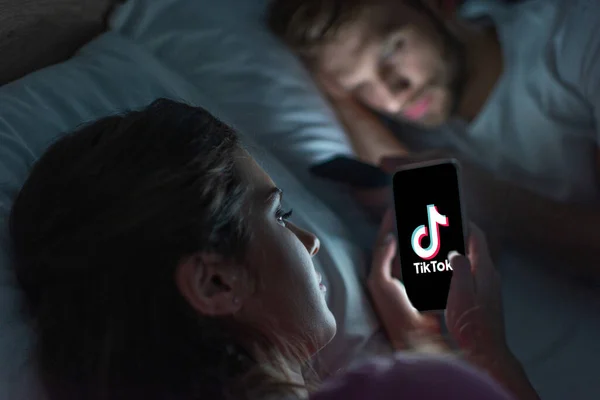 KYIV, UKRAINE - 21 FÉVRIER 2020 : Mise au point sélective d'une jeune femme tenant un smartphone avec l'application TikTok près du petit ami au lit la nuit — Photo de stock