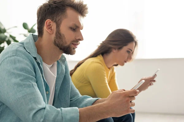 Selective focus of man using smartphone near girlfriend chatting in living room — Stock Photo