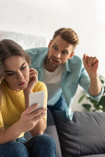 Enfoque selectivo de la chica usando el teléfono inteligente cerca de hombre enojado en la sala de estar - foto de stock