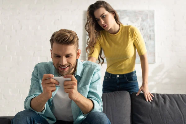 Selective focus of smiling smartphone depended man sitting on couch near girlfriend in living room — Stock Photo