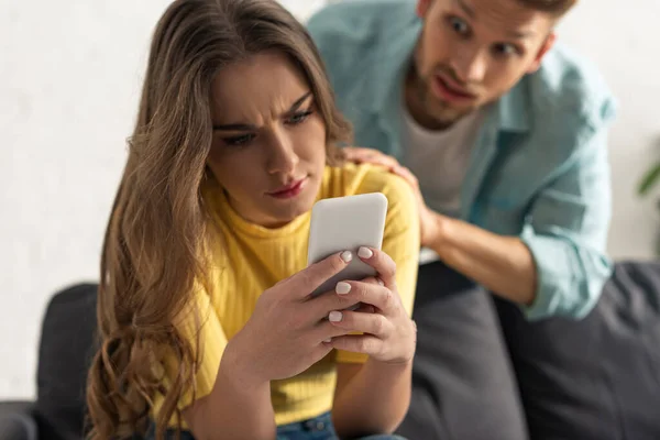 Selective focus of confused girl using smartphone near angry boyfriend in living room — Stock Photo