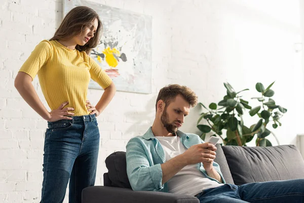 Mujer enojada con las manos en las caderas de pie cerca de teléfono inteligente dependía del hombre en el sofá - foto de stock