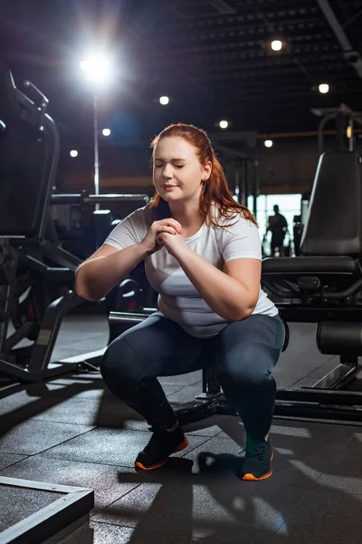 Concentrado chica con sobrepeso con los ojos cerrados en cuclillas con las manos apretadas en el gimnasio — Stock Photo