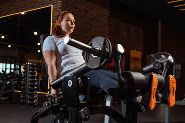Bonita, chica con sobrepeso haciendo ejercicio de extensión de pierna en la máquina de entrenamiento - foto de stock