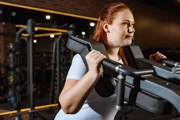 Confident overweight girl doing arms extension exercise on fitness machine — Stock Photo