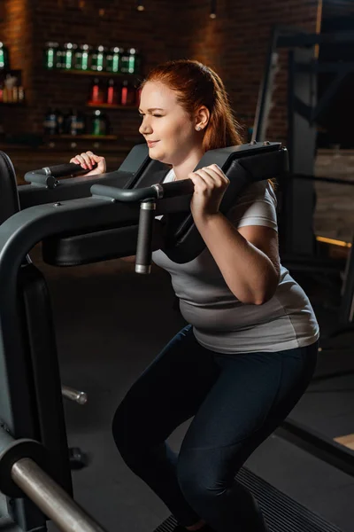 Concentrated overweight girl doing arms extension exercise on fitness machine — Stock Photo