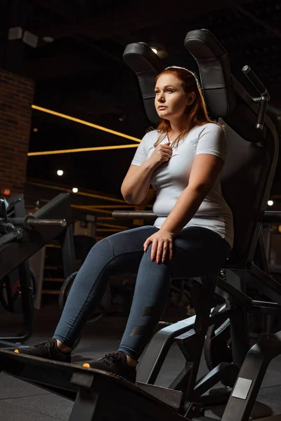 Confident overweight girl looking away while sitting on fitness machine — Stock Photo
