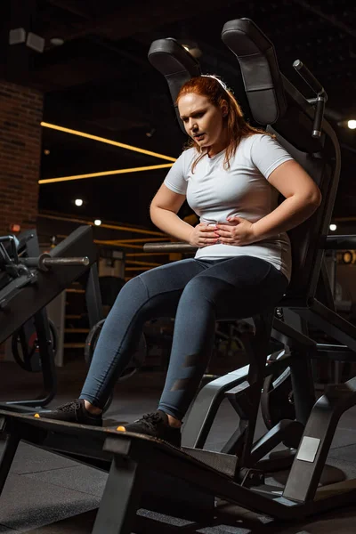 Cansado sobrepeso menina tocando barriga enquanto sentado na máquina de treinamento — Fotografia de Stock