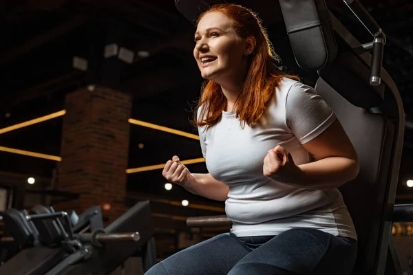 Excited overweight girl showing winner gesture while sitting on fitness machine — Stock Photo