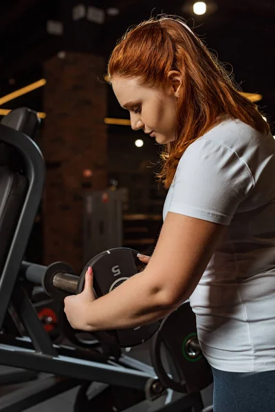 Pretty, overweight girl taking weight disk from fitness machine — Stock Photo