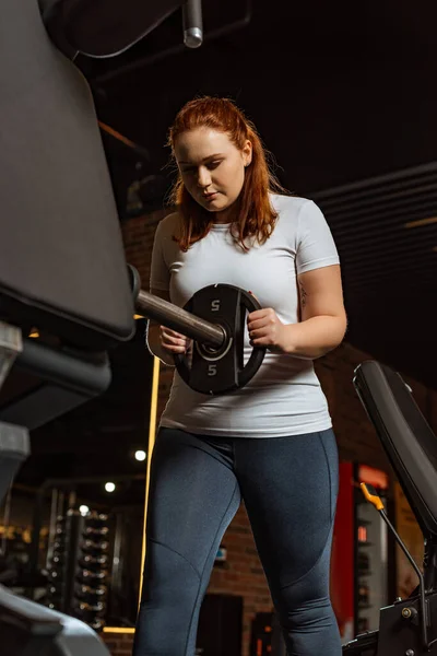 Bonita, chica con sobrepeso tomando disco de peso de la máquina de fitness - foto de stock