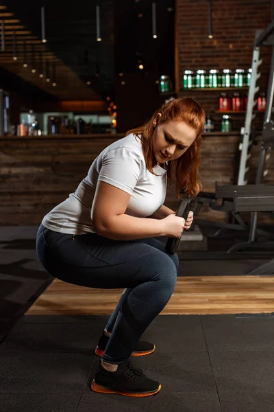 Chica con sobrepeso concentrado en cuclillas con disco de peso en el gimnasio - foto de stock