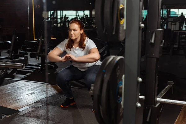 Centrado chica con sobrepeso en cuclillas con las manos apretadas cerca de la máquina de fitness - foto de stock