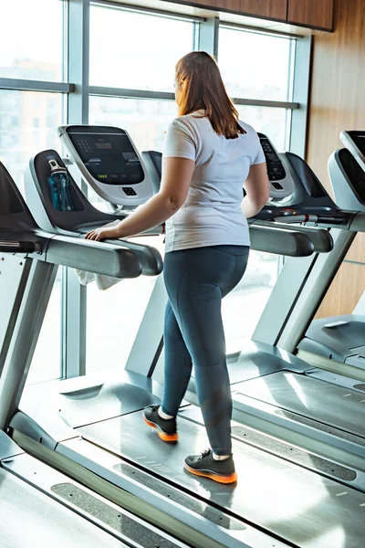 Redhead overweight girl training on treadmill in gym near window — Stock Photo