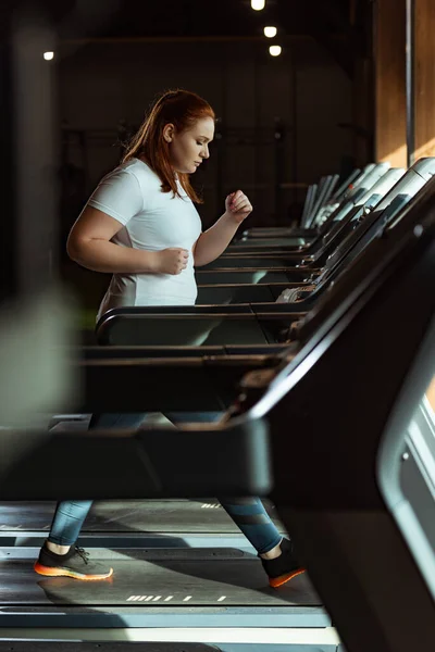 Избирательный фокус сфокусированной девушки с избыточным весом, бегающей на беговой дорожке в спортзале — стоковое фото