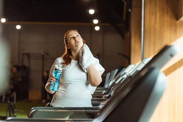 Enfoque selectivo chica con sobrepeso limpiando el cuello con toalla mientras sostiene la botella de deportes - foto de stock