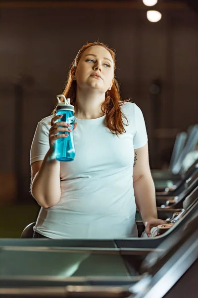 Cansada chica con sobrepeso sosteniendo botella deportiva mientras está de pie en la cinta de correr en el gimnasio - foto de stock