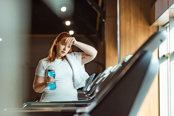 Selective focus of tired overweight girl touching forehead and holding sports bottle while standing at treadmill — Stock Photo