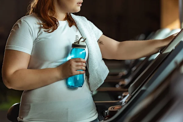 Vista recortada de niña con sobrepeso con toalla en el hombro sosteniendo botella deportiva mientras está de pie en la cinta de correr - foto de stock