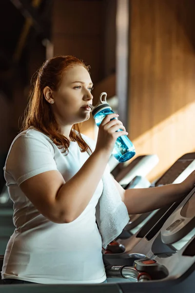 Pensive overweight girl drinking from sports bottle while standing at treadmill — Stock Photo