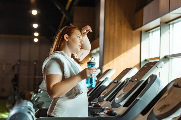 Tired overweight girl touching forehead while holding sports bottle while standing on treadmill — Stock Photo