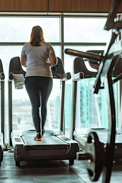 Vista trasera de la chica con sobrepeso corriendo en la cinta de correr en el gimnasio contra la ventana - foto de stock