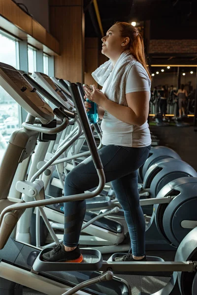 Cansado chica con sobrepeso apertura botella de deportes mientras se entrena en stepper - foto de stock