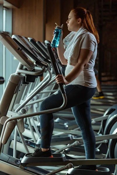 Vista laterale della ragazza in sovrappeso che tiene la bottiglia sportiva mentre si allena sul stepper — Foto stock