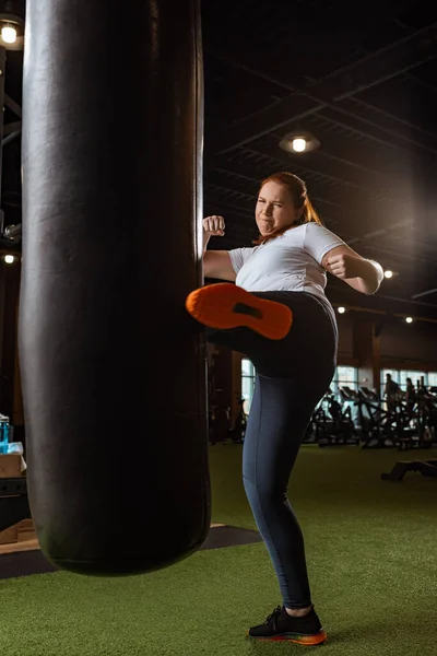 Energizado chica con sobrepeso patadas saco de boxeo con pierna en el gimnasio — Stock Photo