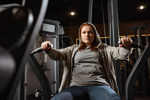 Selective focus of confident overweight girl doing arms extension exercise on fitness machine — Stock Photo