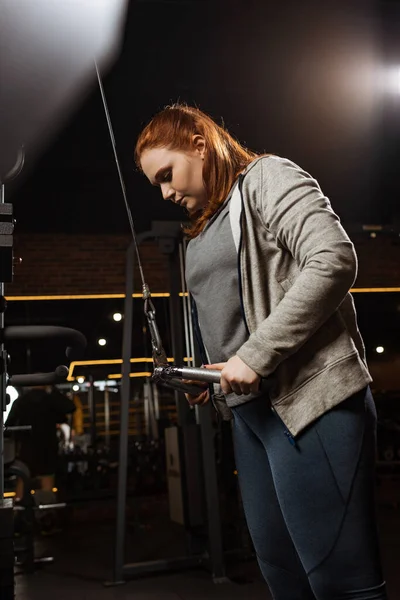 Focused overweight girl doing arms extension exercise on fitness machine — Stock Photo