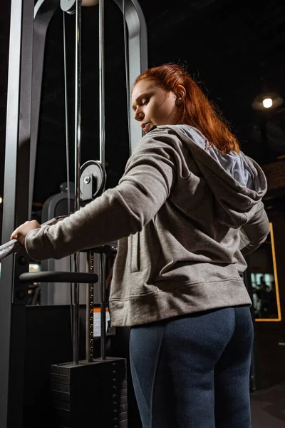 Overweight girl in grey hoodie doing arms extension exercise on fitness machine — Stock Photo