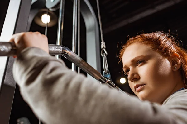 Enfoque selectivo de confianza chica con sobrepeso haciendo ejercicio de extensión de brazos en la máquina de fitness - foto de stock