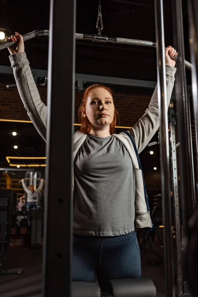 Chica con sobrepeso mirando a la cámara mientras hace ejercicio de extensión de brazos en la máquina de fitness - foto de stock