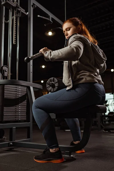 Low angle view of concentrated overweight girl doing arms extension exercise on fitness machine — Stock Photo