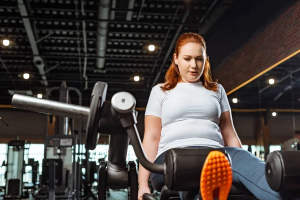 Confiado chica con sobrepeso haciendo ejercicio de extensión de pierna en la máquina de entrenamiento - foto de stock