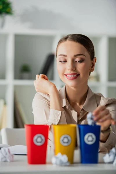 Selective focus of smiling businesswoman throwing crumpled papers into recycling buckets — Stock Photo