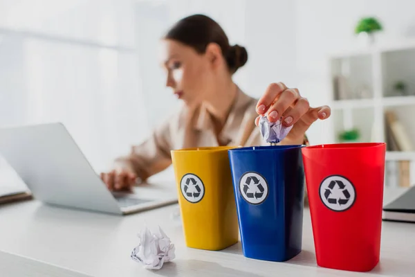 Enfoque selectivo de la mujer de negocios arrojando papeles arrugados en cubos de reciclaje mientras trabaja en el ordenador portátil - foto de stock