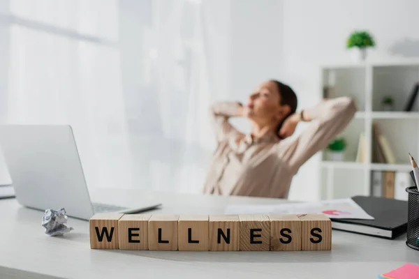 Selective focus of businesswoman relaxing at workplace with laptop and alphabet cubes with wellness word — Stock Photo