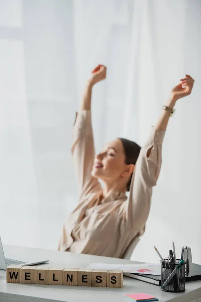 Selective focus of happy businesswoman resting at workplace with alphabet cubes with wellness word — Stock Photo