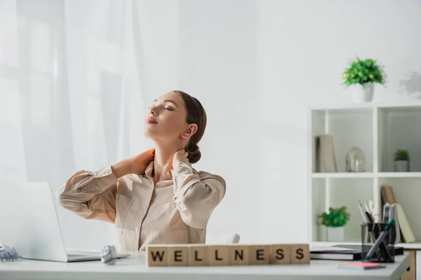 Attractive businesswoman relaxing at workplace with laptop and alphabet cubes with wellness word — Stock Photo