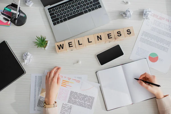 Cropped view of businesswoman writing and working in office with documents, laptop, smartphone and alphabet cubes with wellness word — Stock Photo