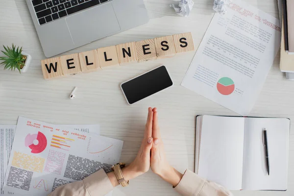 Vista recortada de la mujer de negocios haciendo Namaste gesto en el lugar de trabajo con documentos, ordenador portátil, teléfono inteligente y cubos de alfabeto con palabra de bienestar - foto de stock