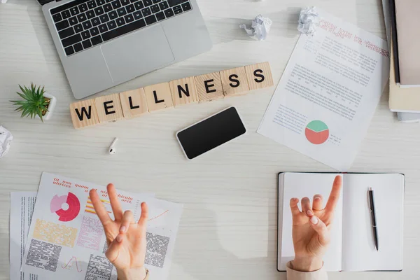 Cropped view of businesswoman making surya mudra at workplace with documents, laptop, smartphone and alphabet cubes with wellness word — Stock Photo